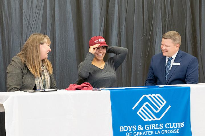 BGC Scholarship Viterbo University President Glena Temple and Boys & Girls Clubs of Greater La Crosse Executive Director Jake Erickson watch as Youth of the Year Awards winner Danessa Brocks tries on a Viterbo hat after signing scholarship papers from the university.
