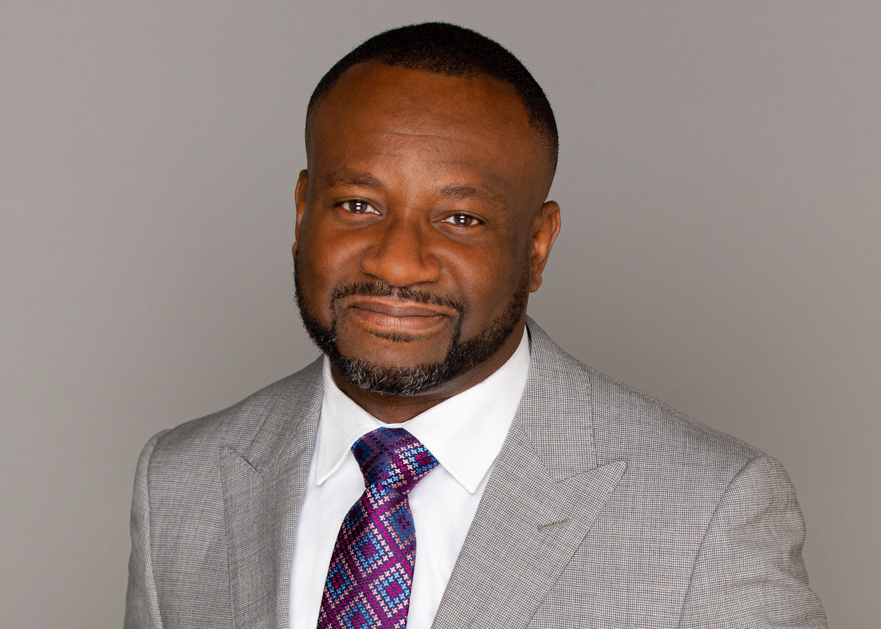 Black man with buzzed hair and a beard in a grey suit with a purple tie