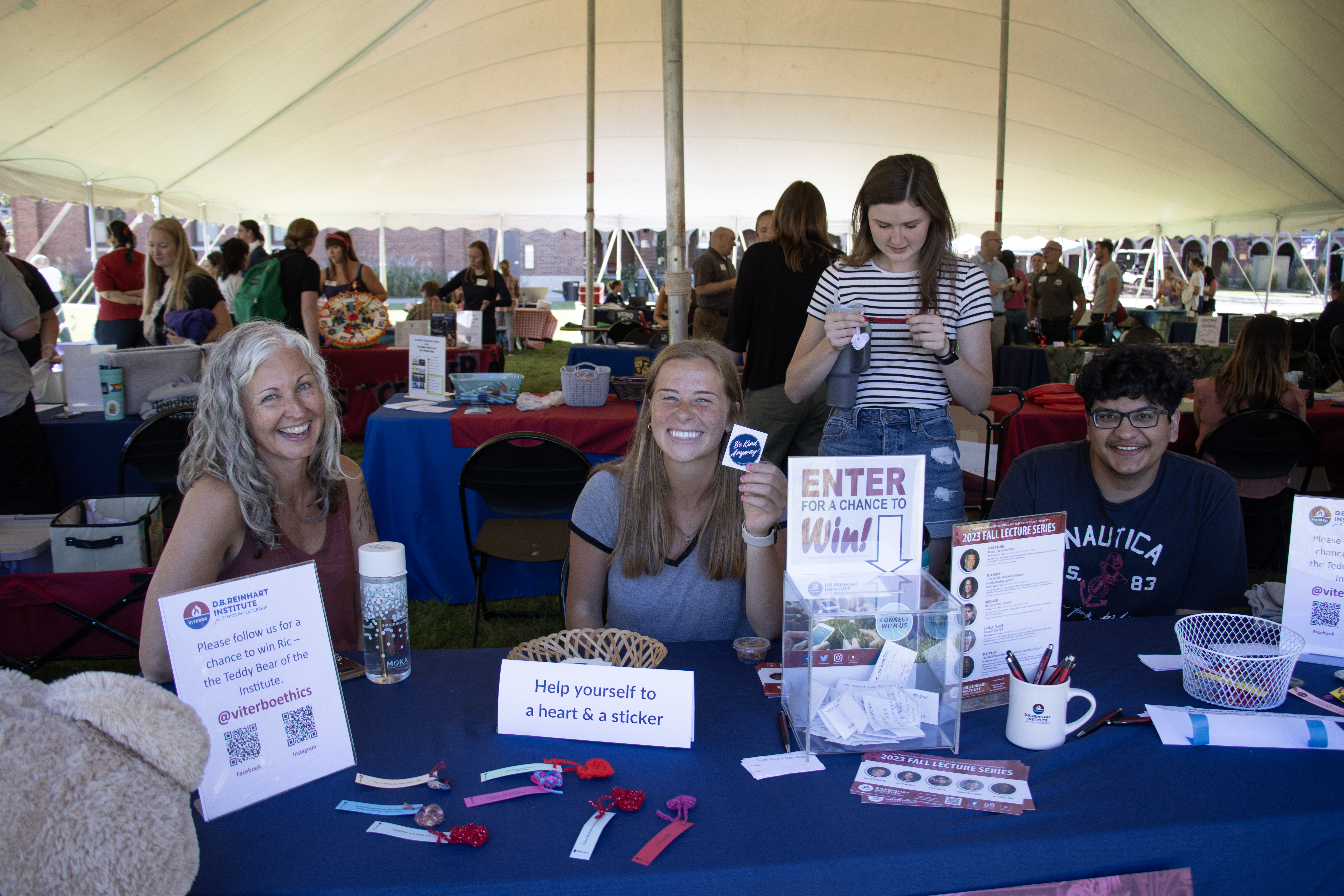 Three members of the D.B. Reinhart institute sit at a table covered in flyers and goodies for Welcome Back Bash. They all smile brightly up at the camera. Behind them, someone stands with a white crochet heart. 