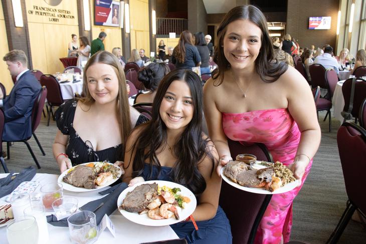 Two women sitting in chairs and one women standing to their right. All three are holding plates of food with beef and shirmp.